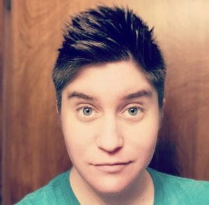 A person with short spiky hair and blue eyes stares directly at the camera, wearing a green shirt. The wooden background adds warmth to the image.