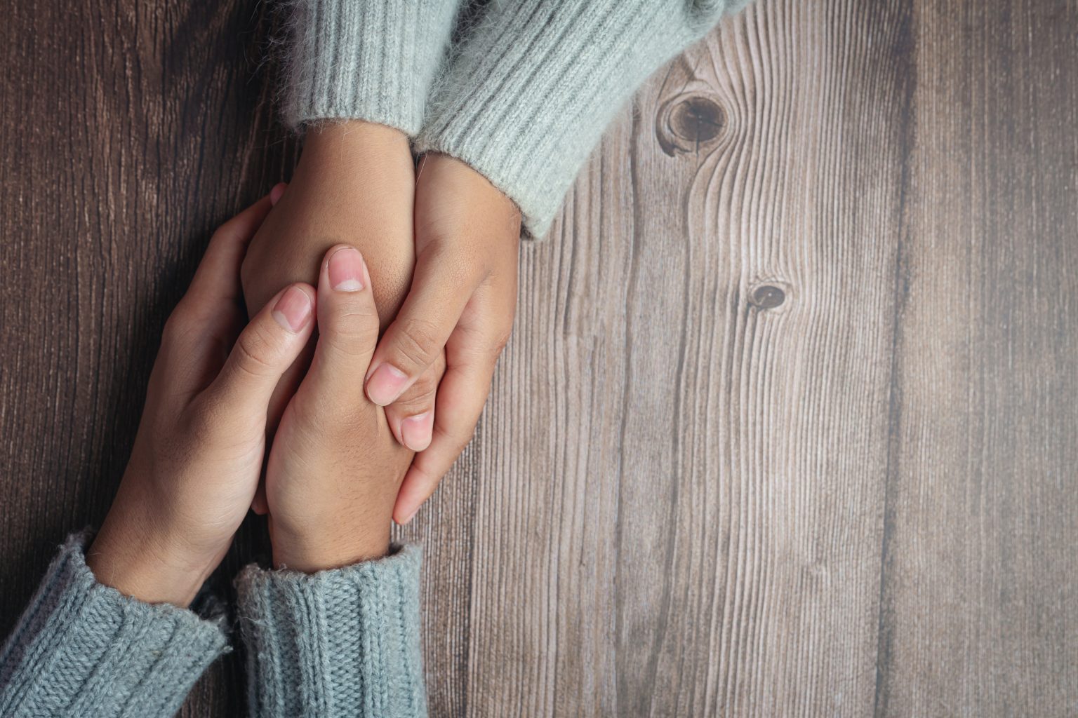 Close-up of two people holding hands on a rustic wooden table, both wearing cozy light gray knit sweaters.